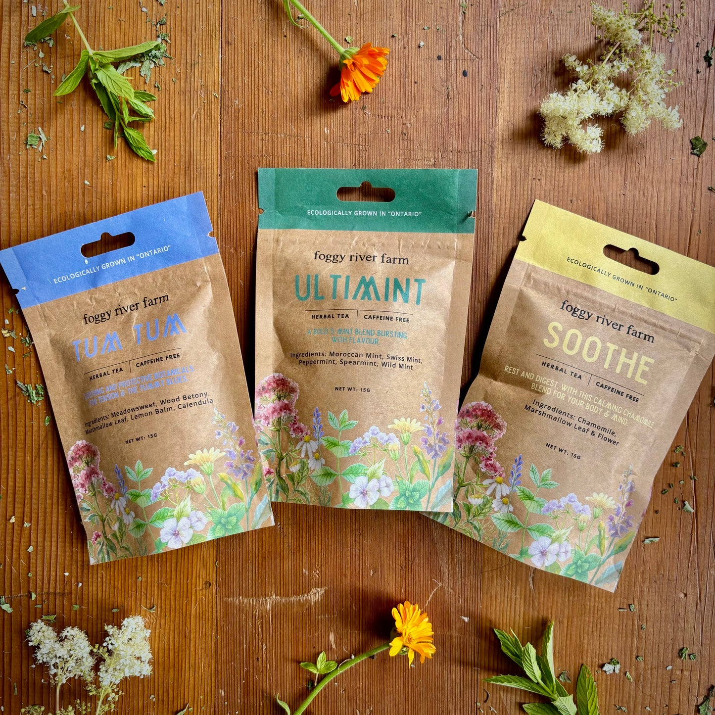 Three packages of foggy river farm herbal teas labeled Soothe, Ultimint, and Tum Tum, displayed on a wooden surface surrounded by fresh herbs and flowers.
