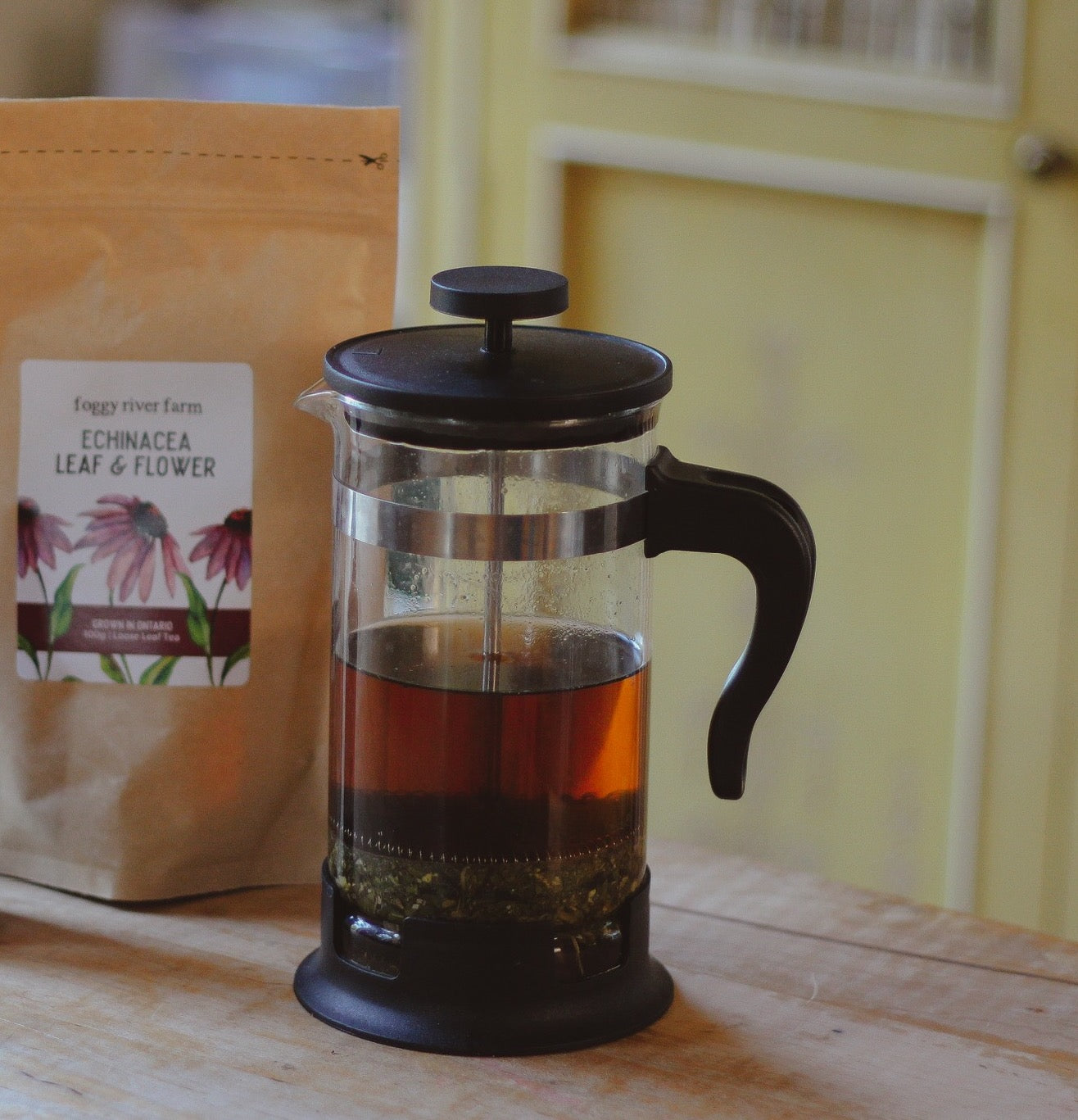 A glass French press filled with brewed echinacea tea sits on a wooden table next to a paper pouch of Echinacea Leaf & Flower from Enggy River Farm, with a softly blurred yellow interior background.