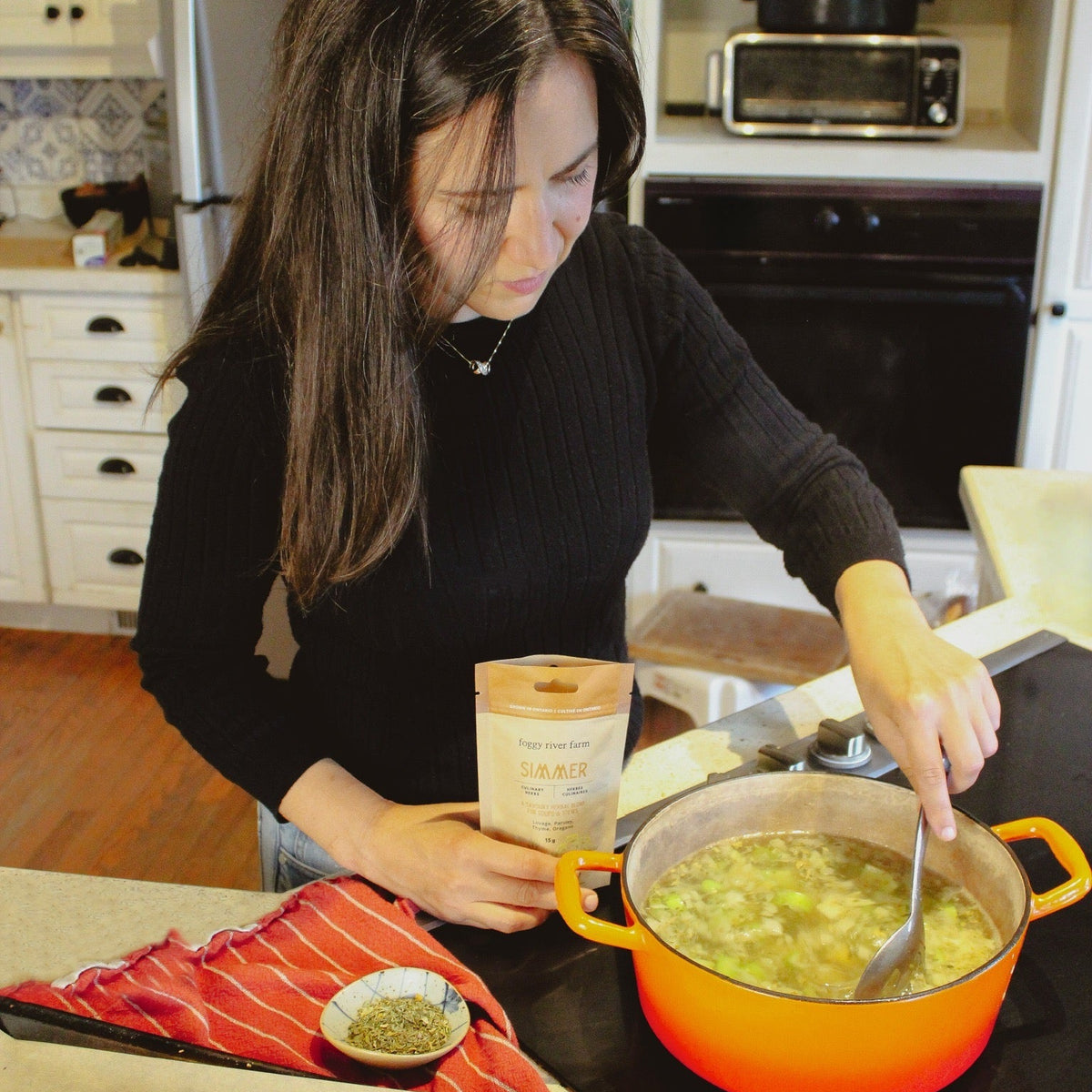 A woman in a black long-sleeve top stands in a kitchen, stirring a green soup in a bright orange pot while holding a paper bag. A baking tray of roasted potatoes and a small dish of herbs sit on the counter beside her.