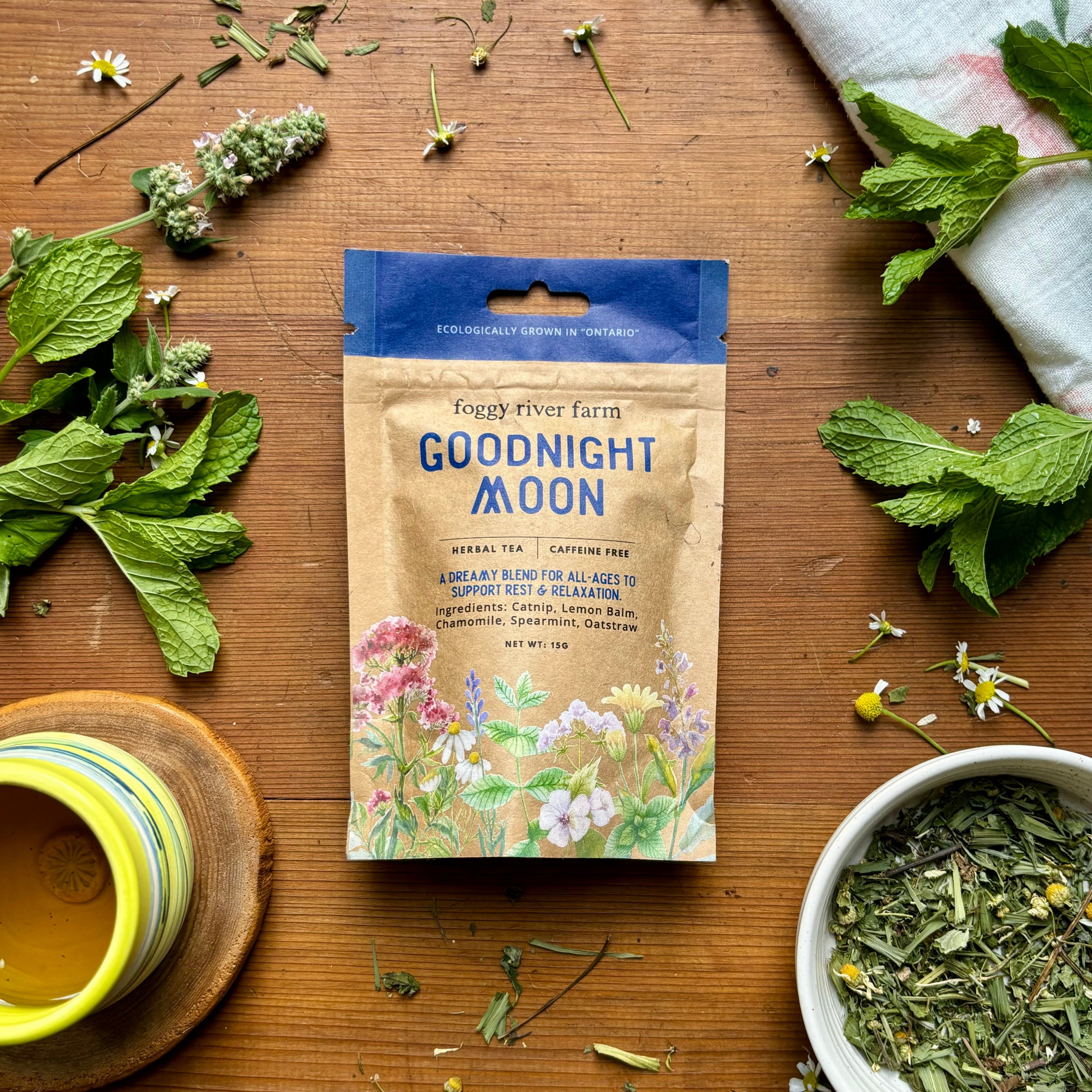 Package of Foggy River Farm's Goodnight Moon Herbal Tea with fresh herbs and flowers displayed around it on a wooden surface