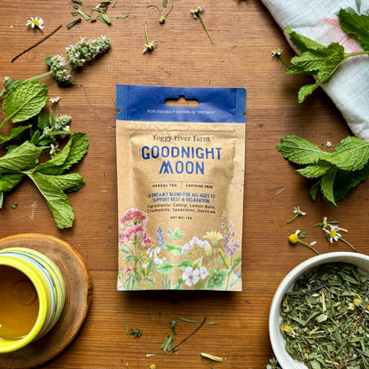Package of Foggy River Farm's Goodnight Moon Herbal Tea with fresh herbs and flowers displayed around it on a wooden surface