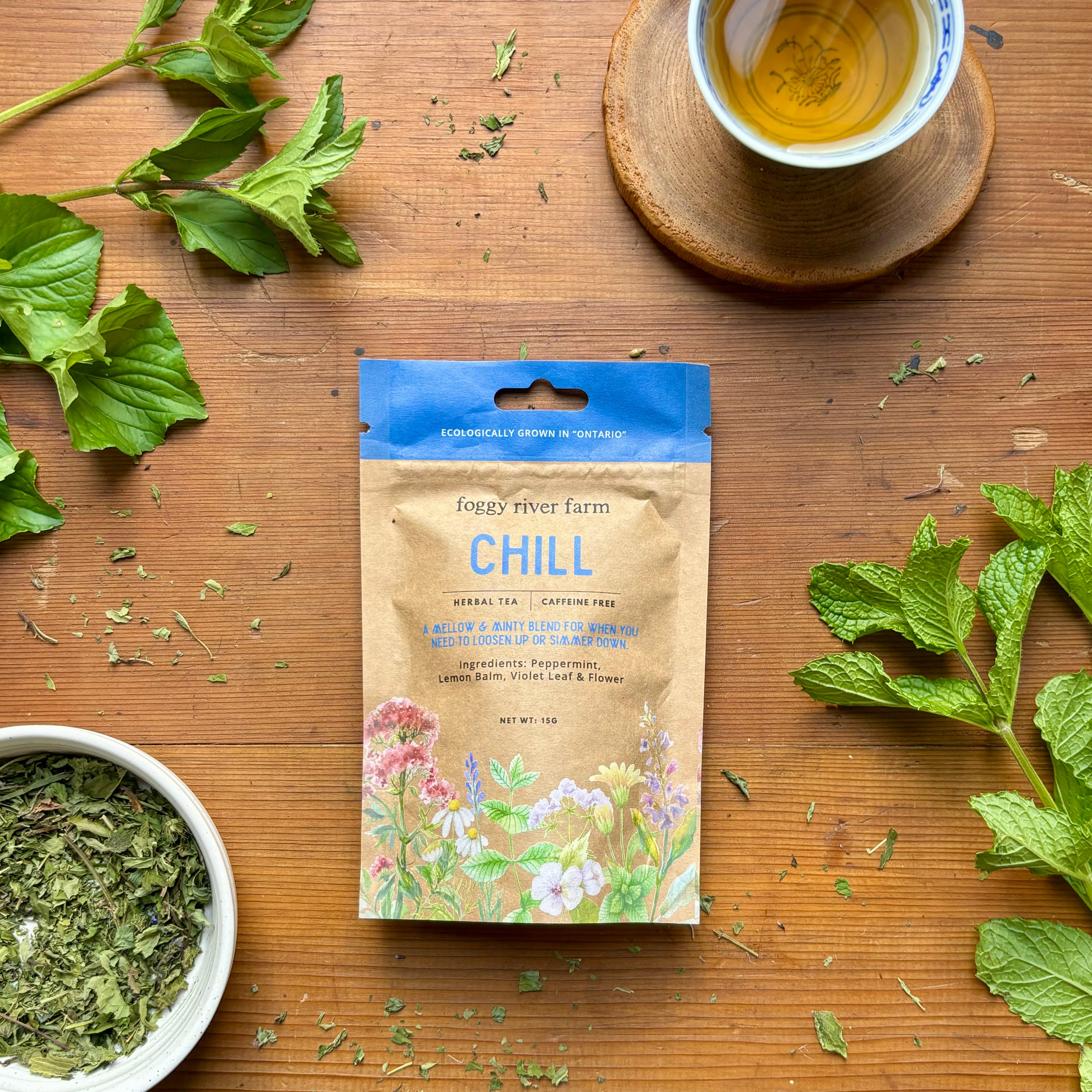 Package of Foggy River Farm's Chill Herbal Tea displayed with fresh herbs and a cup of tea on a wooden surface background