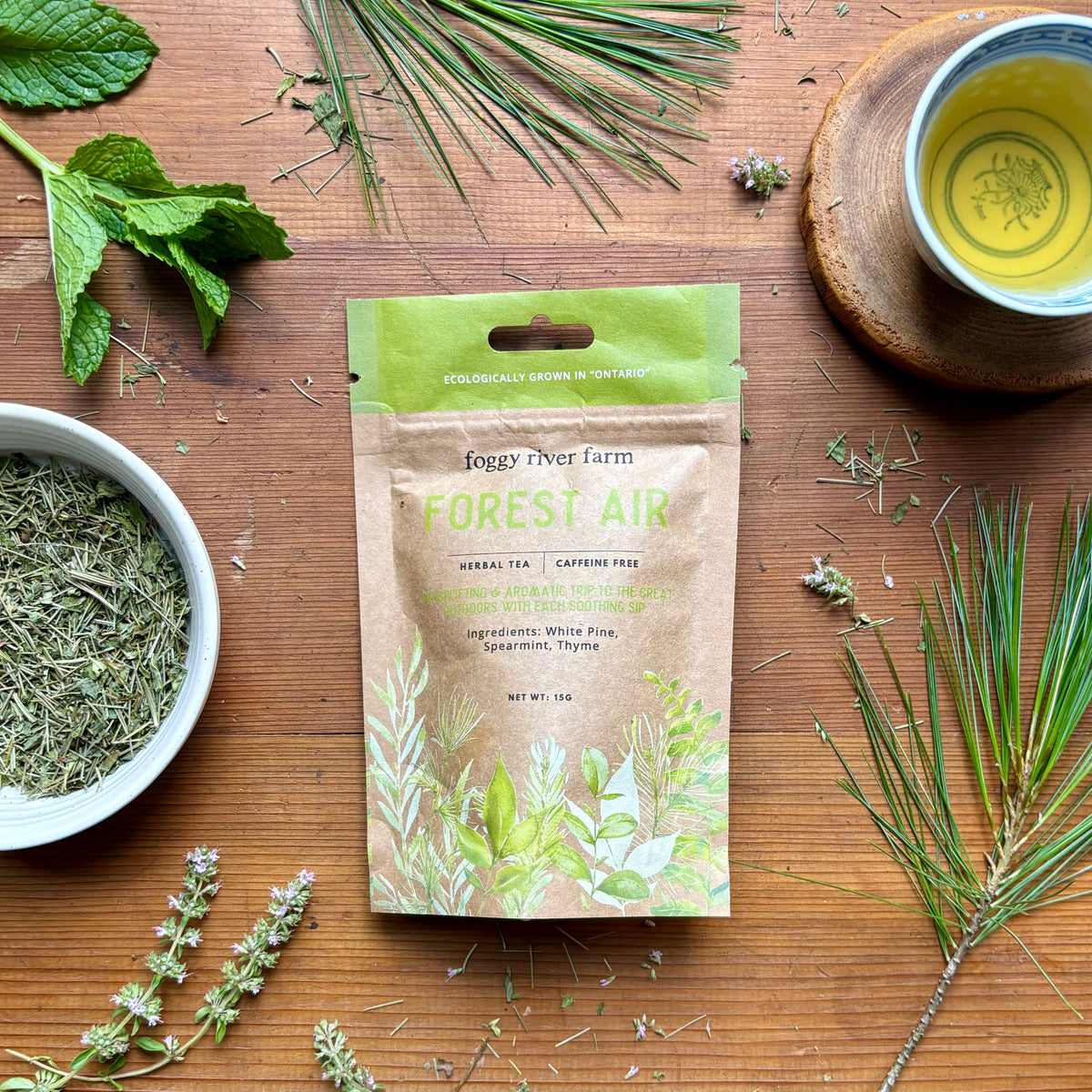 Craft brown package of foggy river farm's forest air herbal tea surrounded by fresh herbs and a cup of tea on a wooden background.  