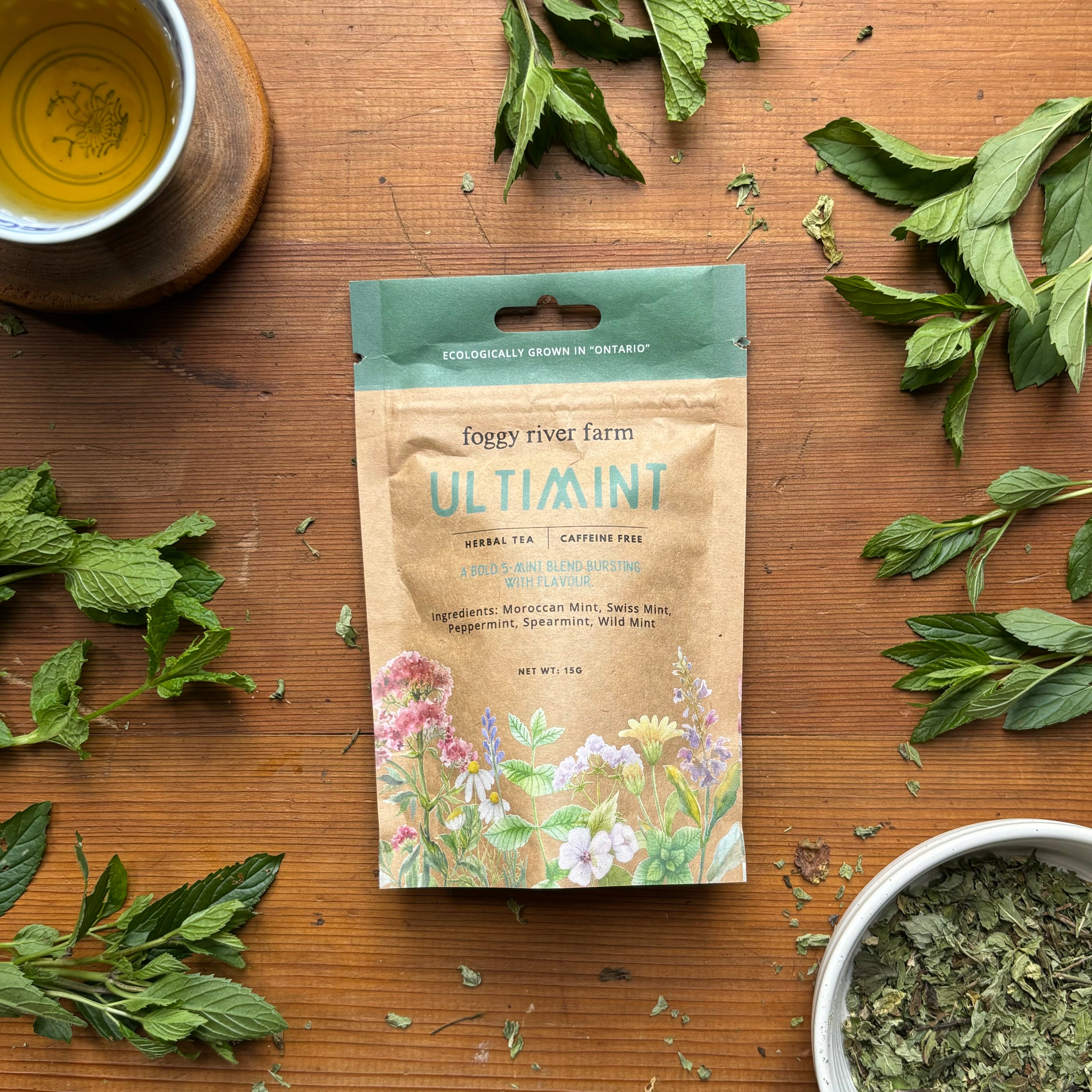 Brown craft package of foggy river farm's ultimint herbal tea surrounded by fresh mint on a wooden surface.