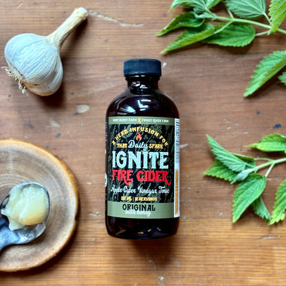 A bottle of Ignite Fire Cider surrounded by a garlic clove, a lemon slice, and some green herbs on a wooden surface