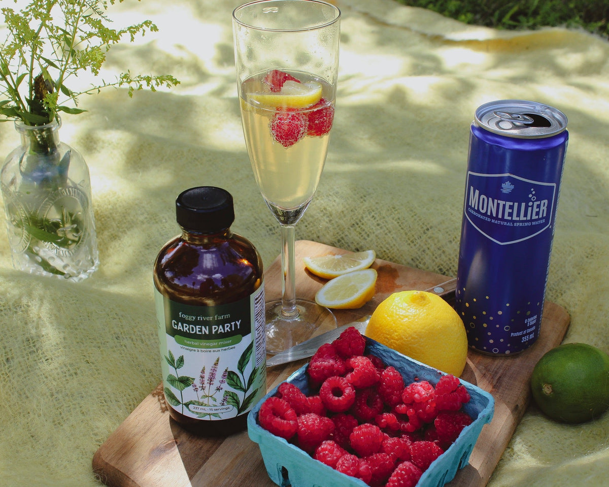 A styled outdoor picnic scene on a yellow blanket with a glass of sparkling drink topped with raspberries, a blue Montellier can, a bottle labeled Garden Party syrup, a tray of fresh raspberries, lemons, limes, and a small vase of flowers.